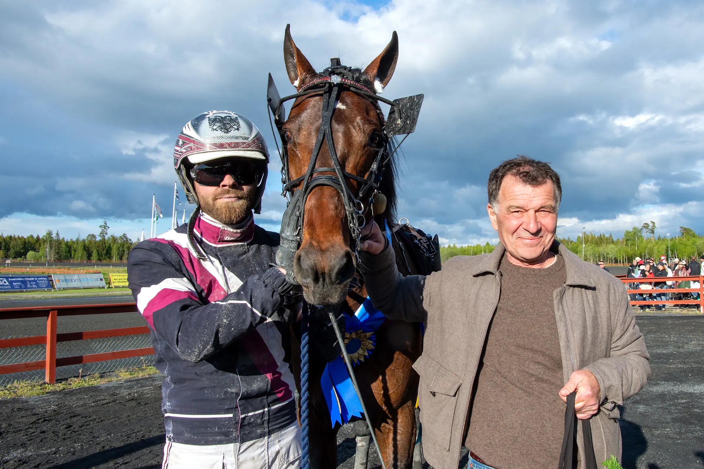 Jockey i färgglad tävlingsuniform och en man i beige jacka står tillsammans med en brun racinghäst på en travbana. I bakgrunden syns läktare och träningsanläggningar under molnig himmel.