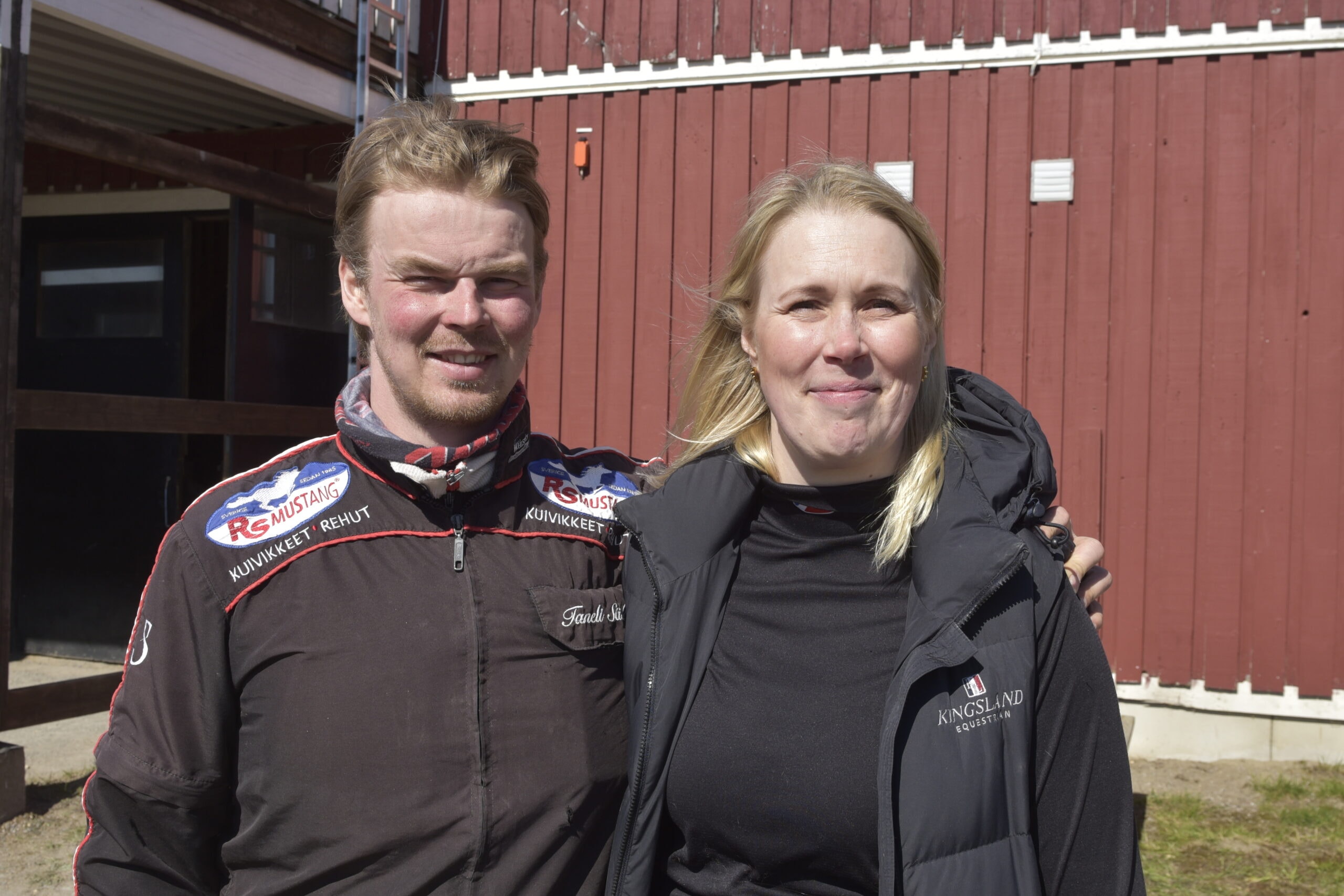 Två personer står tillsammans och ler mot kameran framför en röd träbyggnad. Personen till vänster bär en mörkbrun jacka med märken, och personen till höger bär en svart vinterjacka.