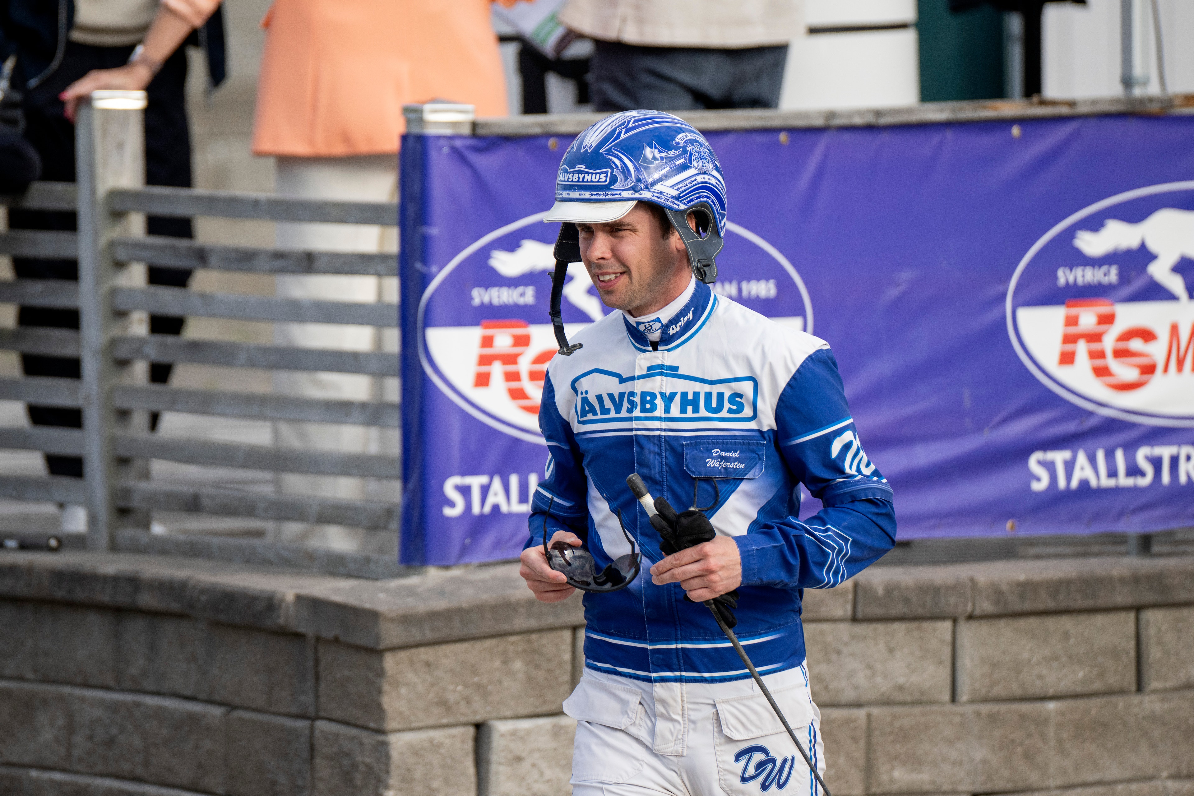 En racerförare i blå och vit racingdräkt och blå hjälm står med två trofeer i händerna. I bakgrunden syns sponsorbanderoller och åskådare.