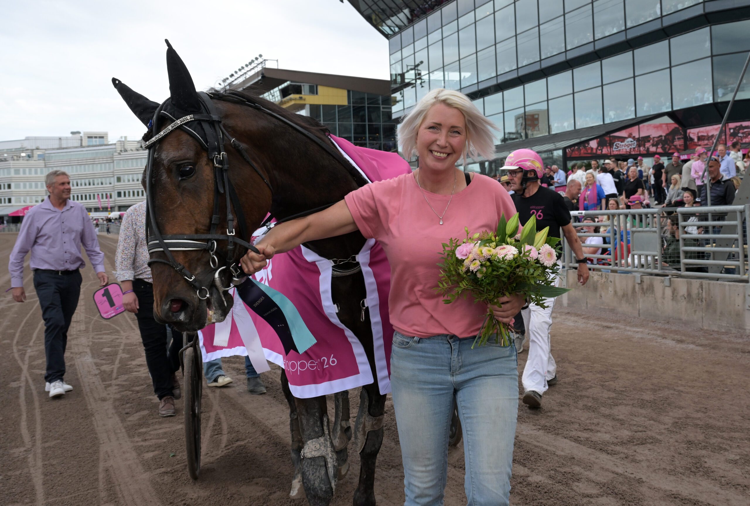 En kvinna i rosa skjorta står bredvid en brun häst på en sandplan vid ett tävlingsarrangemang. Kvinnan håller hästen i tygeln och håller en bukett blommor. I bakgrunden syns publik, moderna byggnader och tävlingsanläggningar.