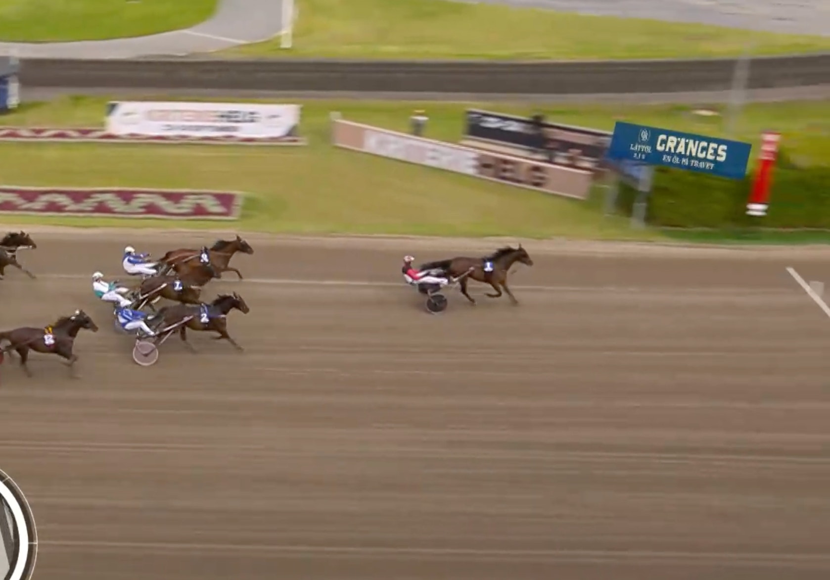 Travhästar i full galopp under ett kapplöpningslopp på en bana med gräs och sponsorskyltar i bakgrunden.