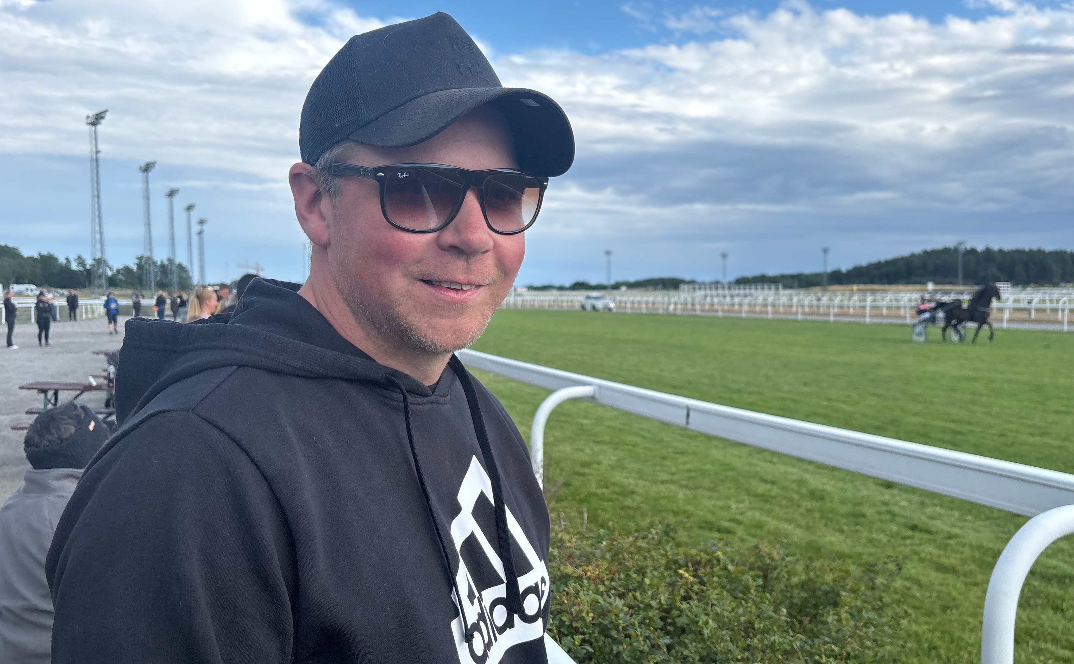 Porträtt av en man vid en travbana. Han bär en svart keps och solglasögon och står framför en vit räcke med travbanan och hästhoppare i bakgrunden.
Foto: Patrick Sjöö