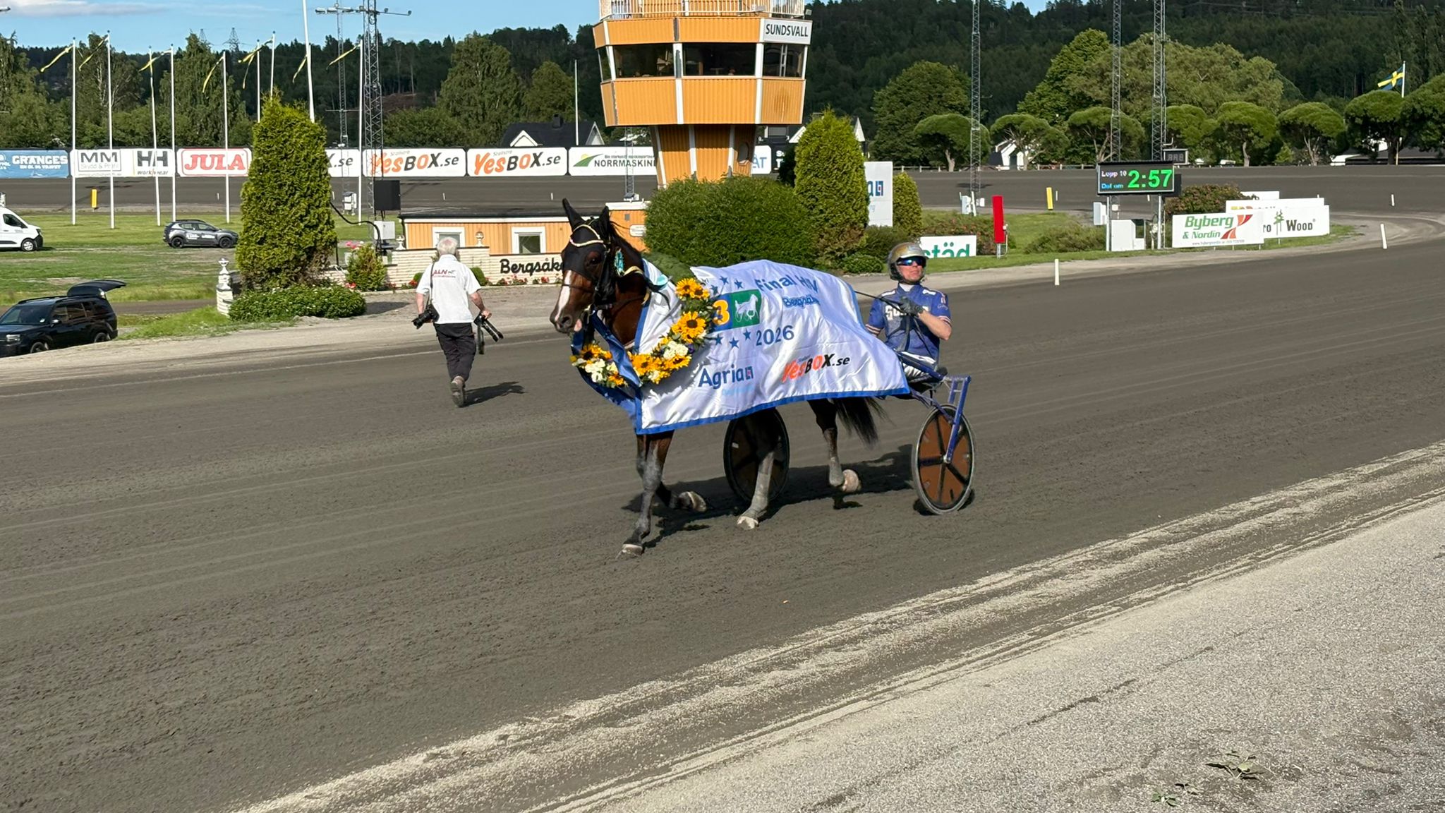 Två personer håller upp en flagga på en racingbana med en orange kontrolltorn i bakgrunden.