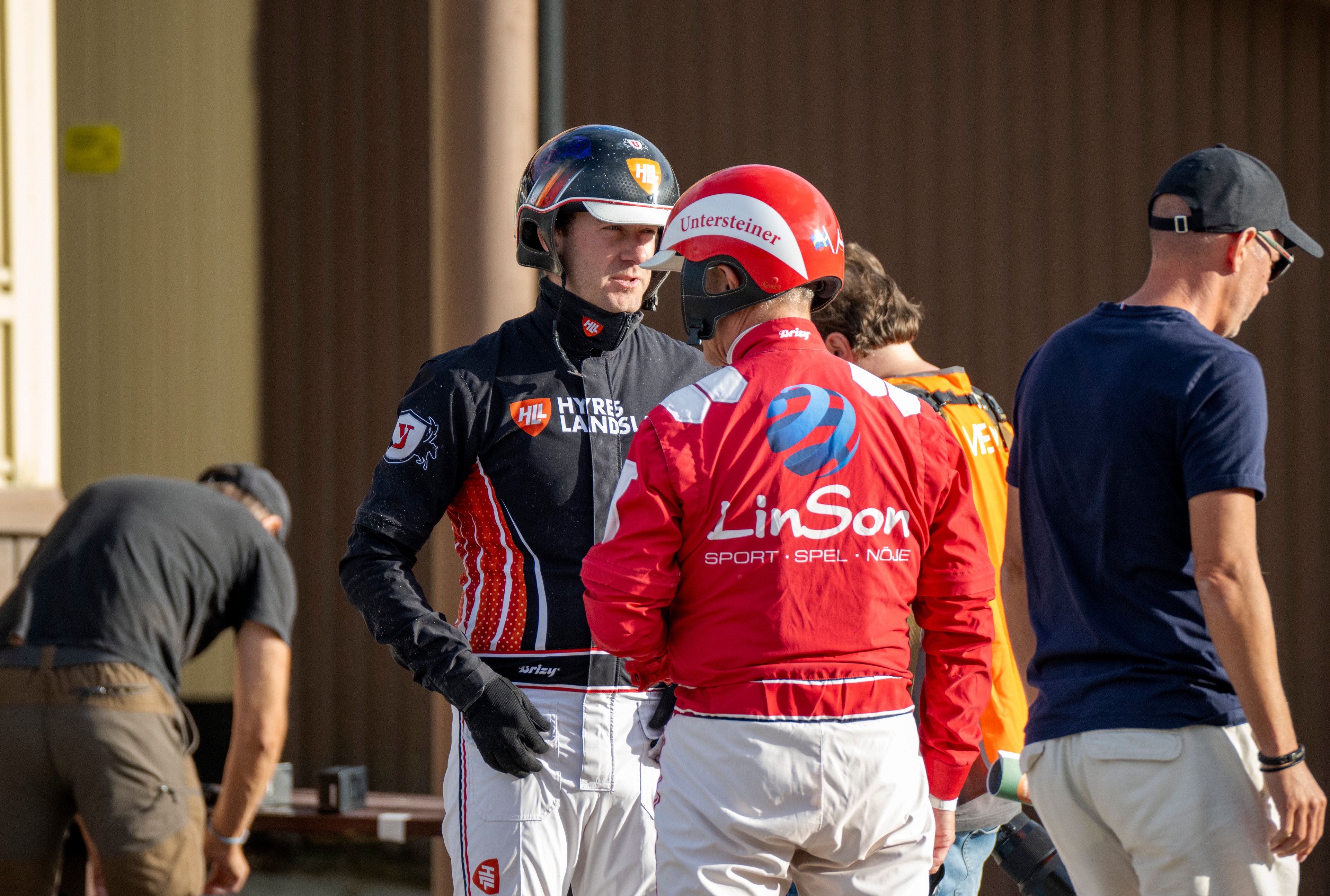 Motorsportförare i racing-utrustning samlade i paddock. En förare i röd overall med LinSon-logotyp står i mitten med hjälm och handskar, omgiven av andra förare och personal.