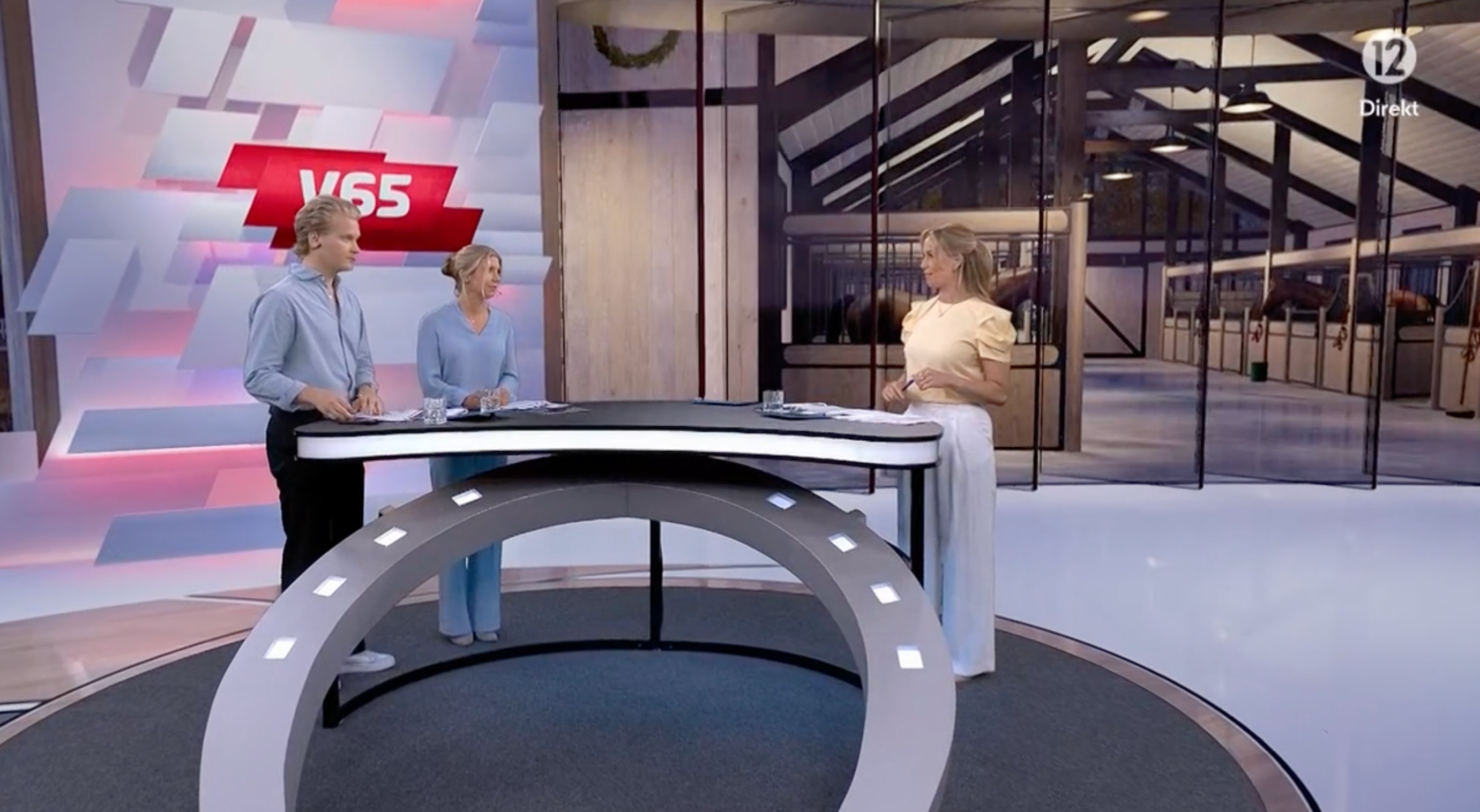 Två personer sitter vid ett modernt nyhetsbord i en TV-studio med V65-logotyp synlig i bakgrunden. En tredje person står i bakgrunden nära glaskontorer.