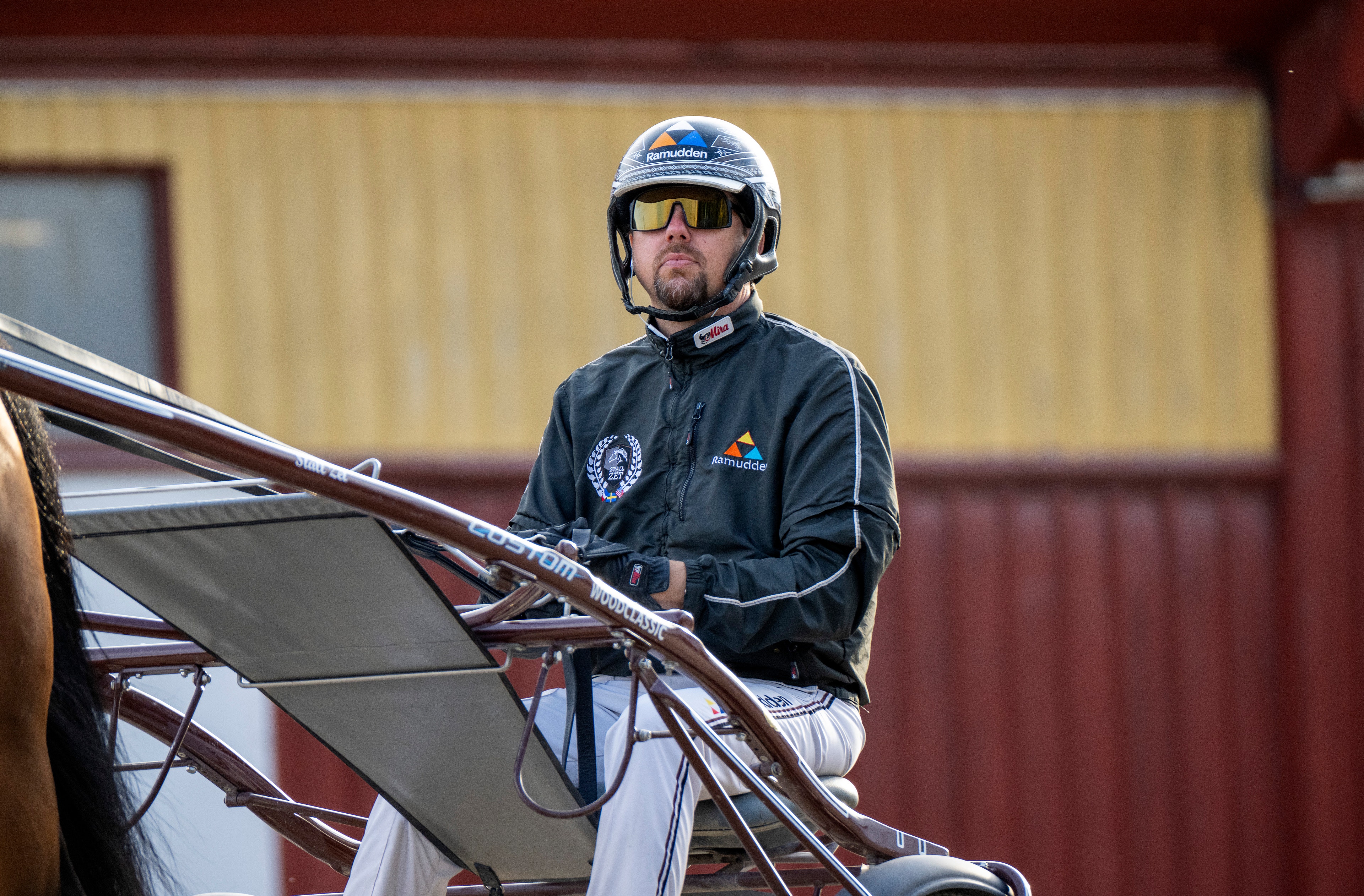 En kuskar i vit hälm och svart jacka sitter i en travvagn bakom en häst under en tävling. I bakgrunden syns en byggnad med röda och gula väggar.