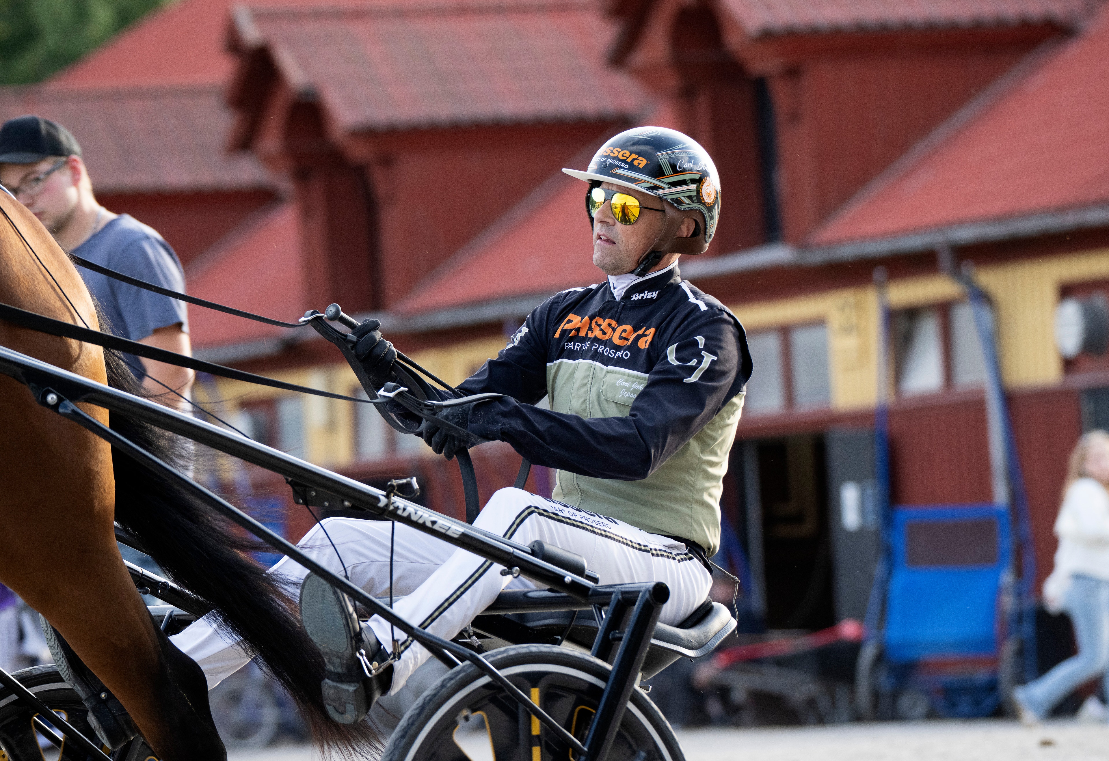 Kusktävling med jockey i svart och vit ridkläder som kör en häst framför rödmålade stallbyggnader.
