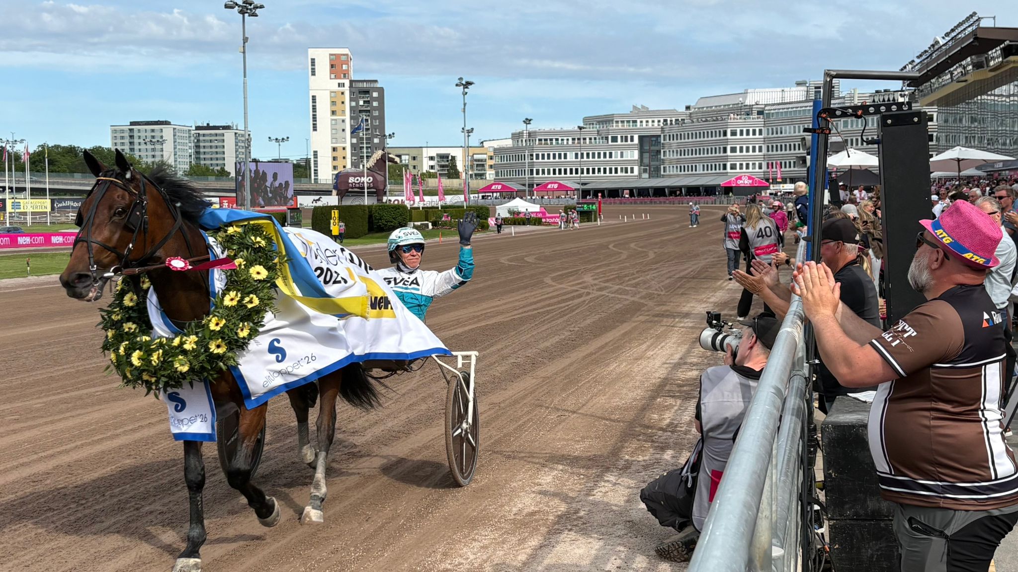 En häst med sulky på en travbana. Hästen är dekorerad i gult och svart. Publik står längs rälerna och fotograferar.