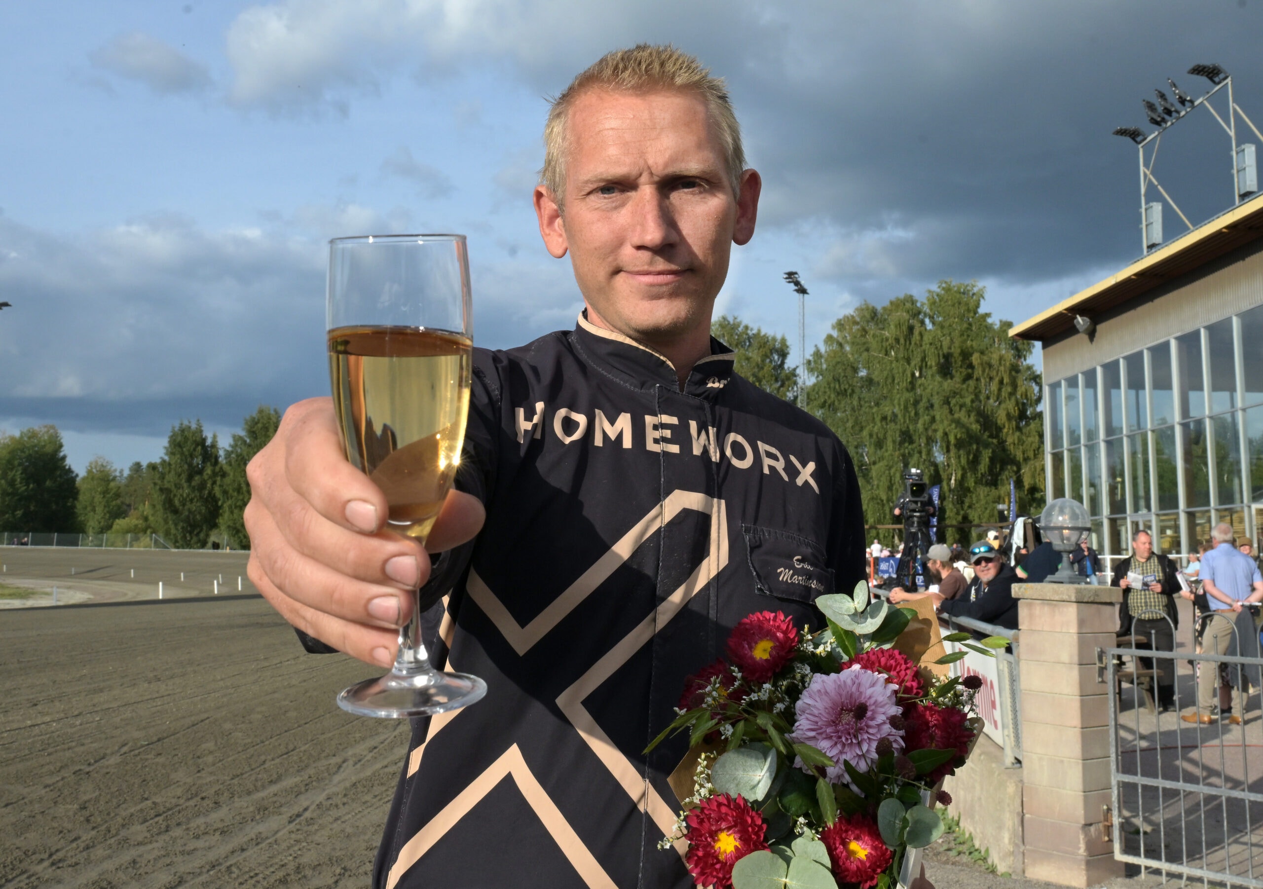 Man i svart tävlingsdräkt håller upp ett champagneglas och en blomsterbukett på en motorsportbana för att fira en seger.