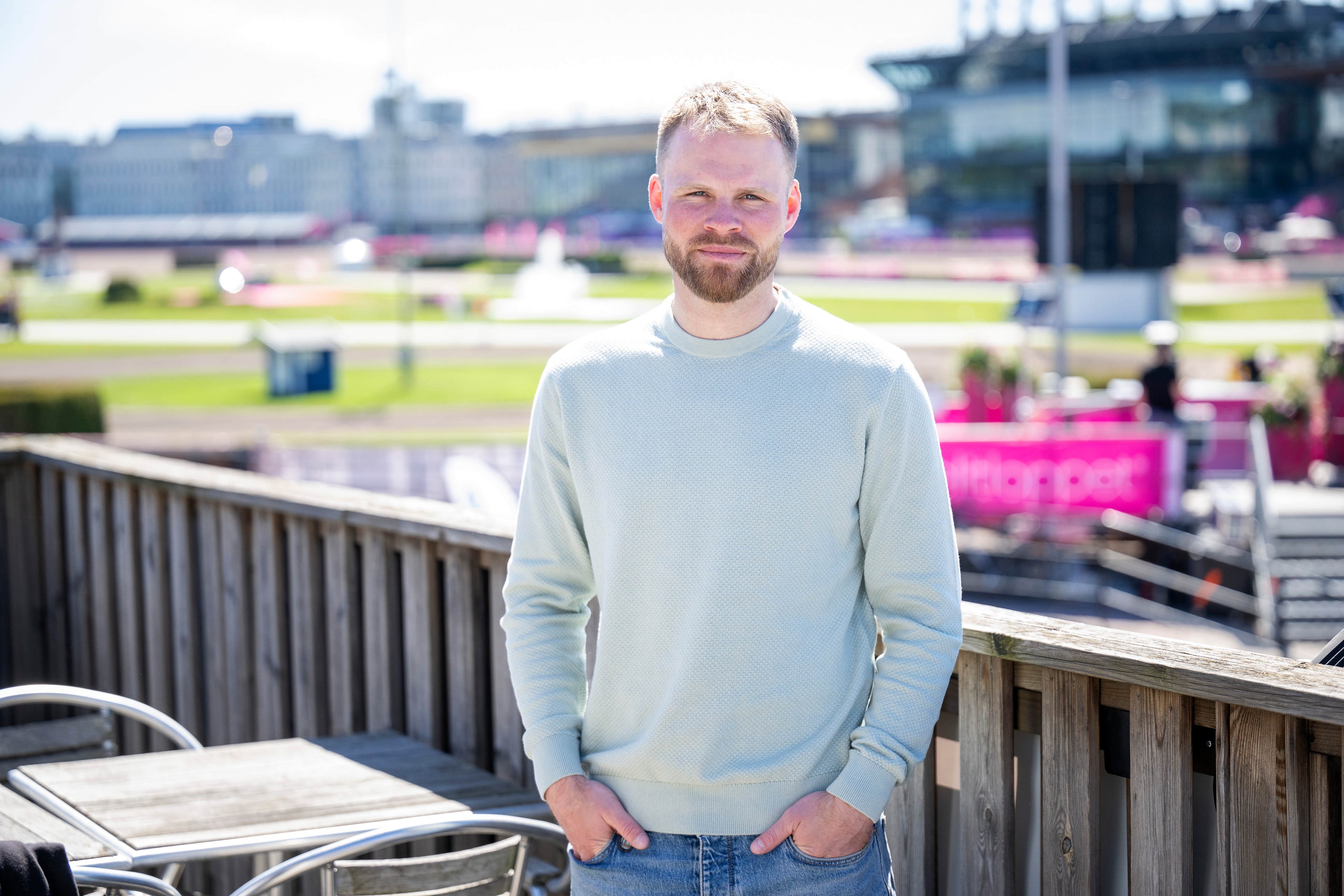 En man i ljusblå sweatshirt står på en balkong med utsikt över Solvalla travbana. I bakgrunden syns gräsplan, läktare och byggnadskomplex.