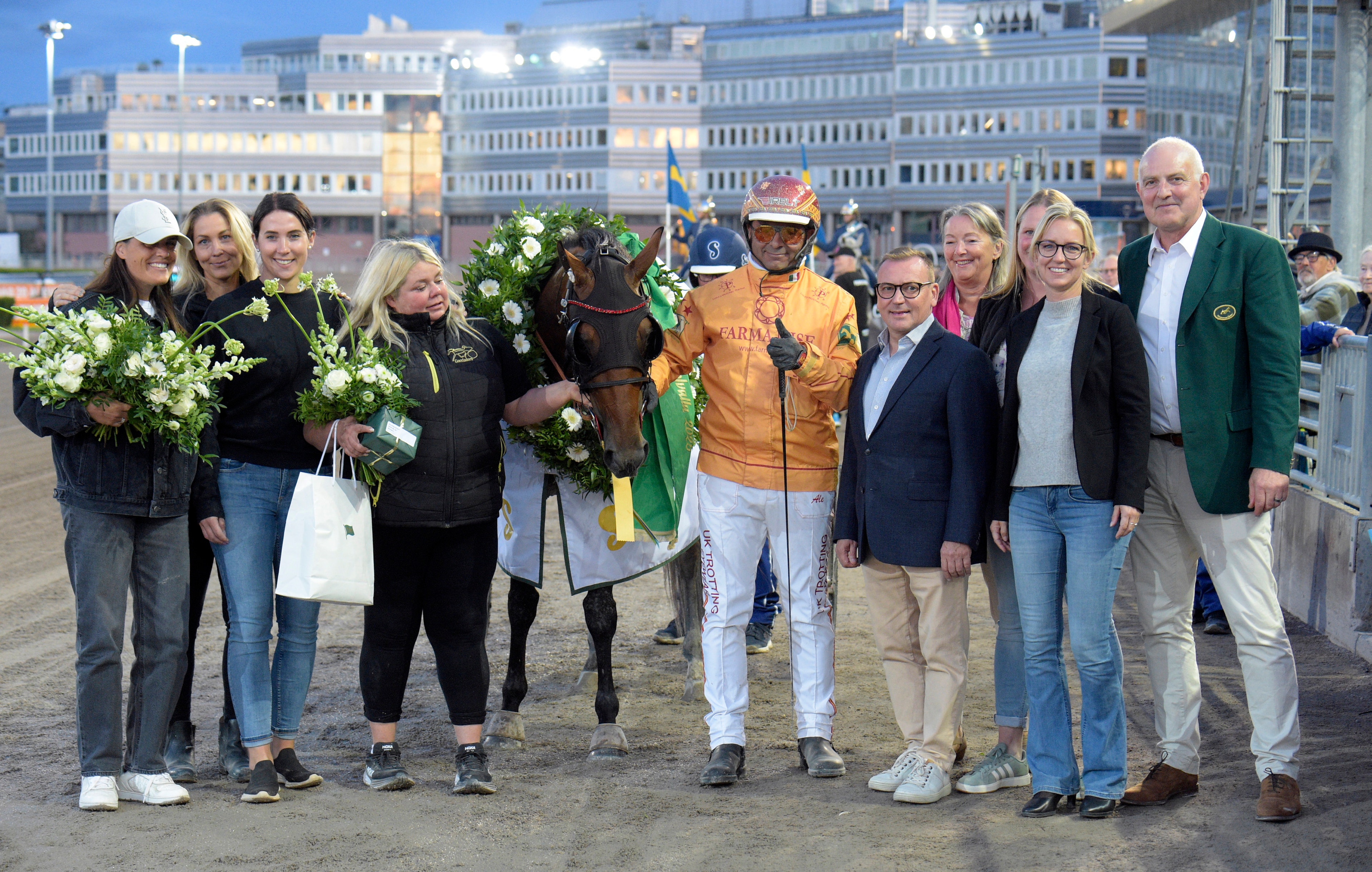Gruppfoto med flera personer som står tillsammans på en öppen plats framför höghus. En person i gul jockey-jacka står bredvid en häst, och flera av personerna håller vita blommor. Det verkar vara en officiell eller festlig sammankomst.