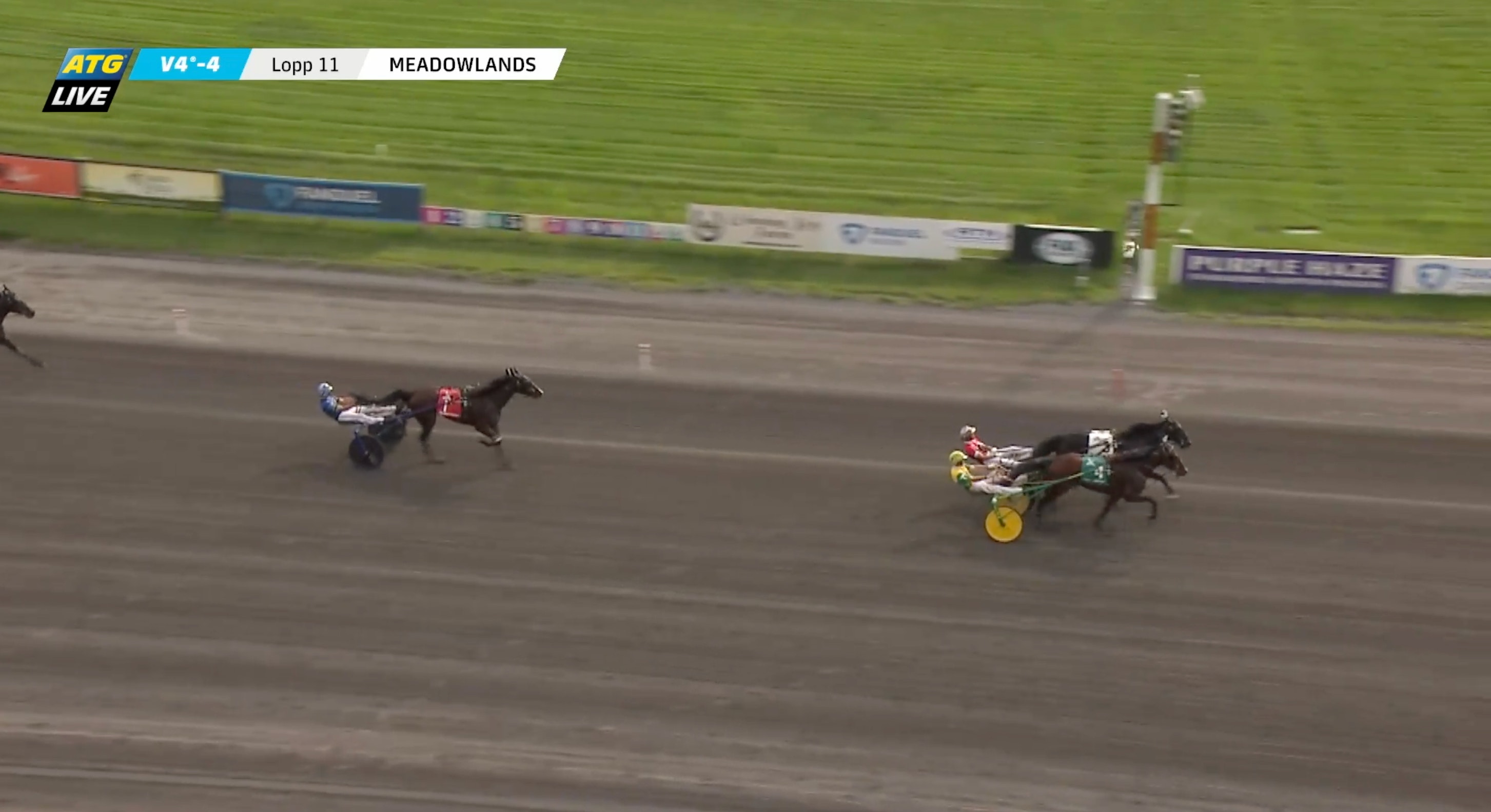 Hästkapplöpning på Meadowlands-banan med två travhästar som tävlar på en sandtrack med gröna gräsområden och sponsorbanderoller längs sidorna.