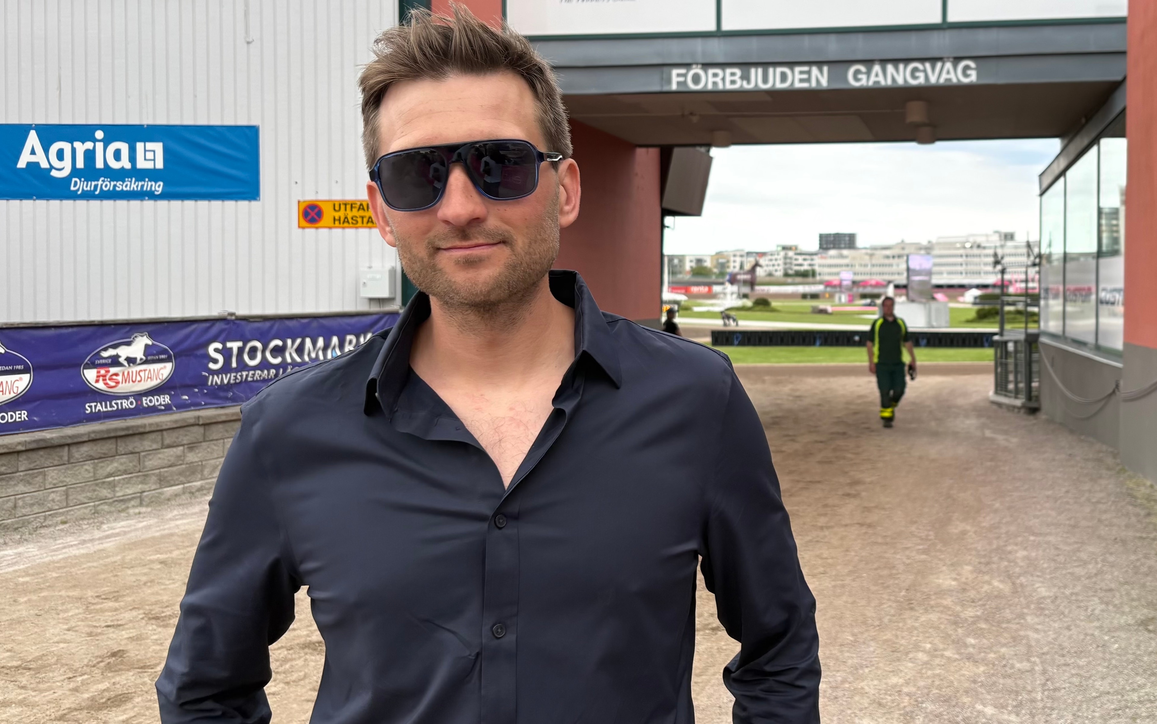 En man med mörka solglasögon och mörkblå skjorta står utomhus på en grusplan vid en byggnad. I bakgrunden syns företagslogotyper och en grön äng.