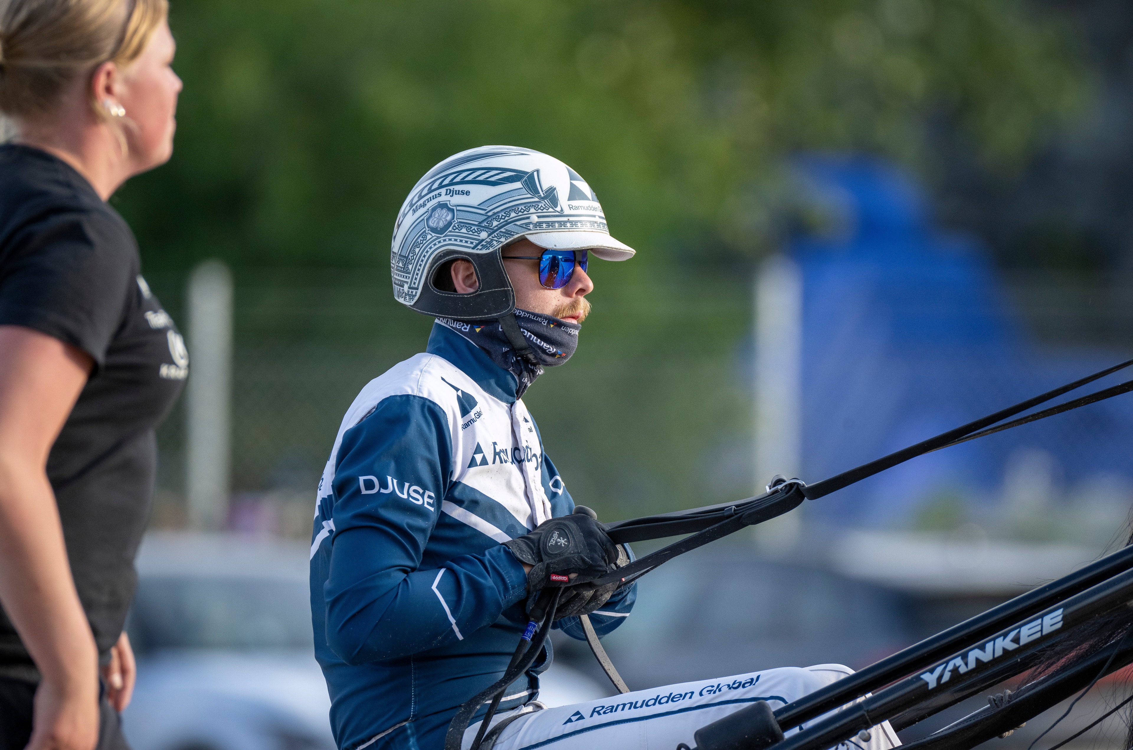 Motionär i blå och vit racingdräkt med hjälm och solglasögon sitter i en båt och håller i ratten. En person i svart skjorta står bredvid. Grön vegetation och blå båtdelar syns i bakgrunden.
Magnus A Djuse