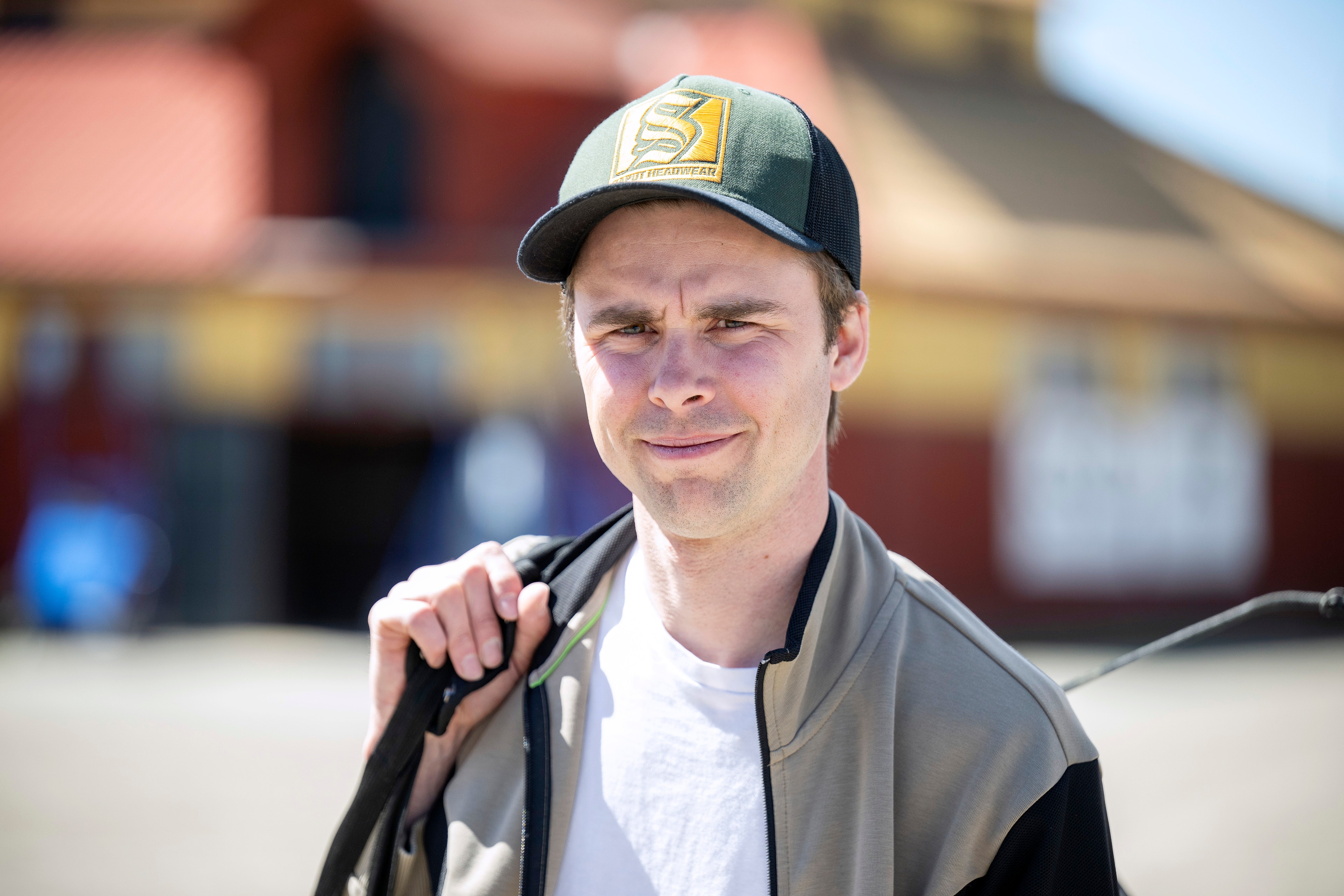 Porträtt av en man som står utomhus framför rödbruna byggnader. Han bär en keps och en jacka över en vit tröja.