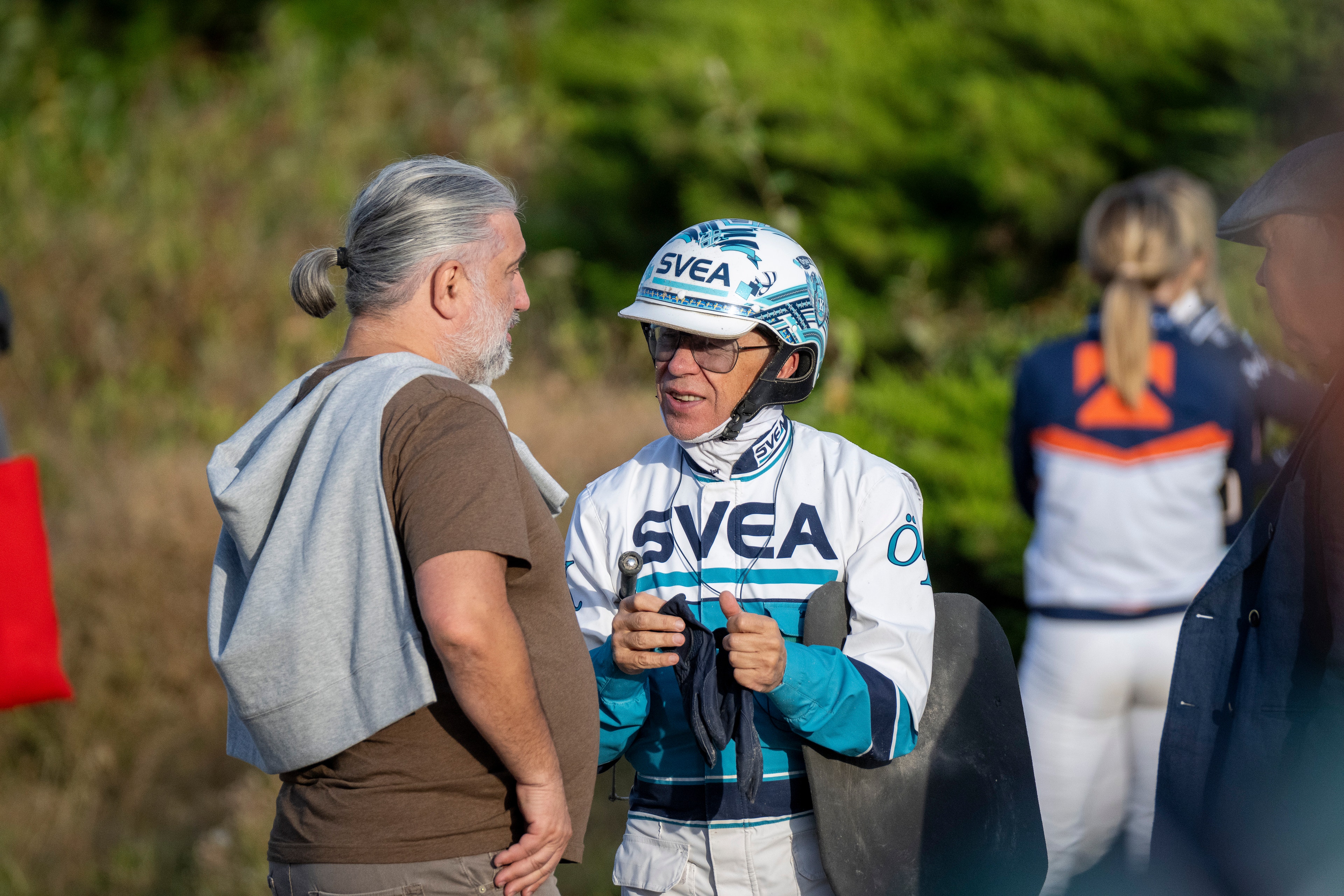 En cyklist i vit och blå SVEA-tröja och cykelhjälm står och samtalar med en man i grå och brun tröja. I bakgrunden syns andra personer och gröna buskar.