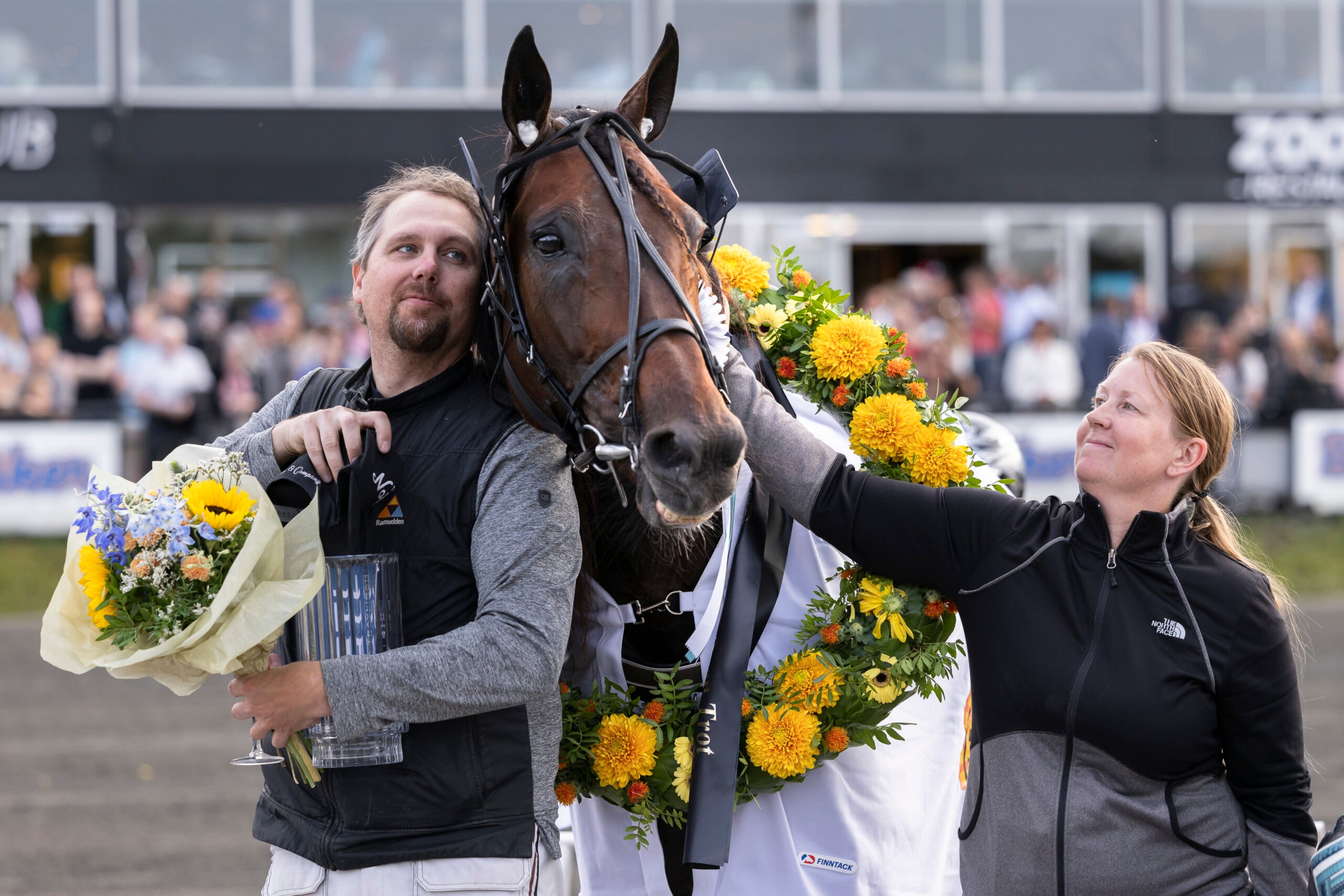 En man och en kvinna står tillsammans med en brun häst som är dekorerad med gula och blå blommor. Mannen håller en blomsterbukett och båda ler. I bakgrunden syns en publik och en byggnad, vilket tyder på ett hästsportevent.
