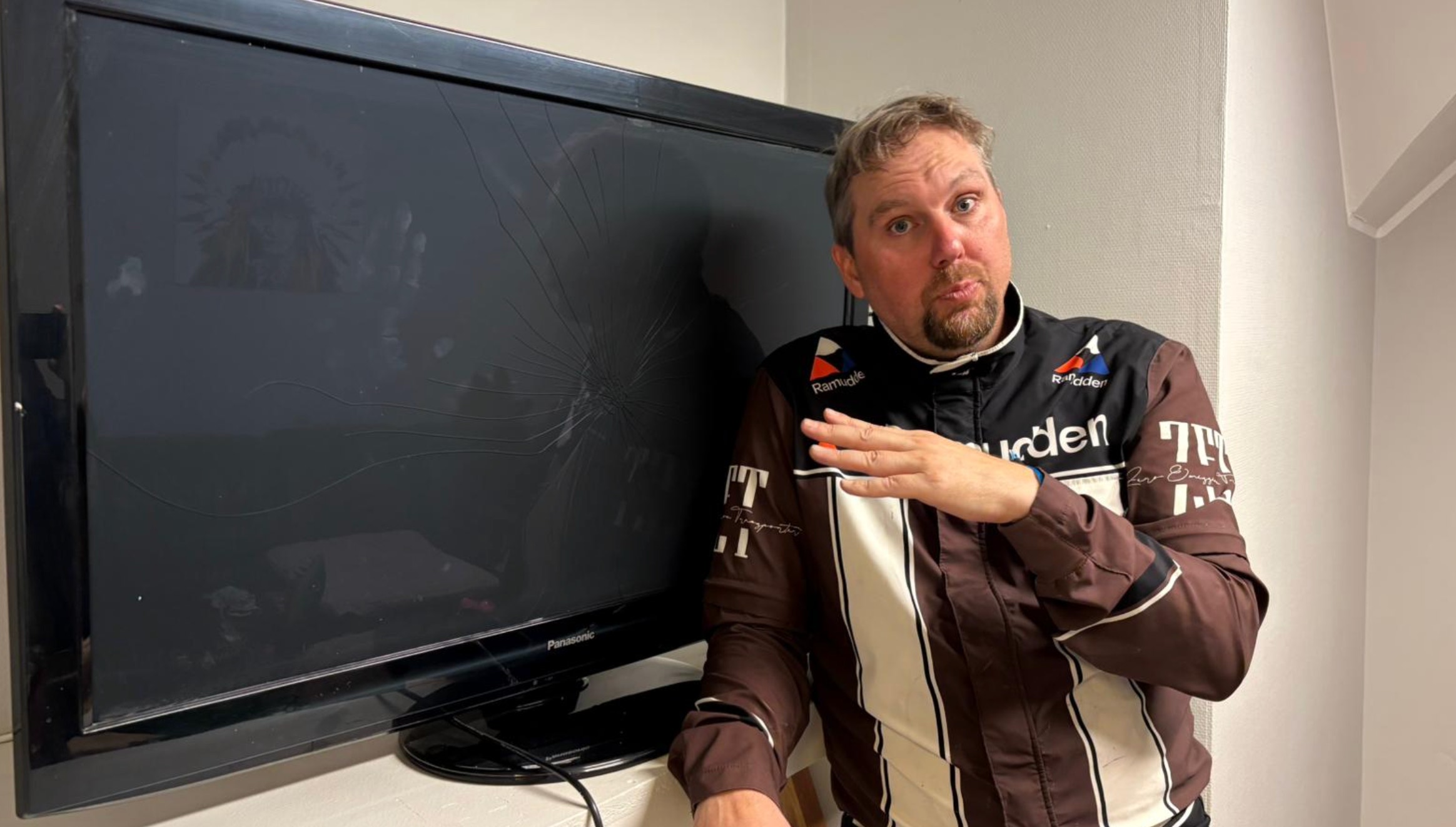 En man i mörkbrun racingoverall står framför en stor TV-skärm och gestikulerar medan han talar.
