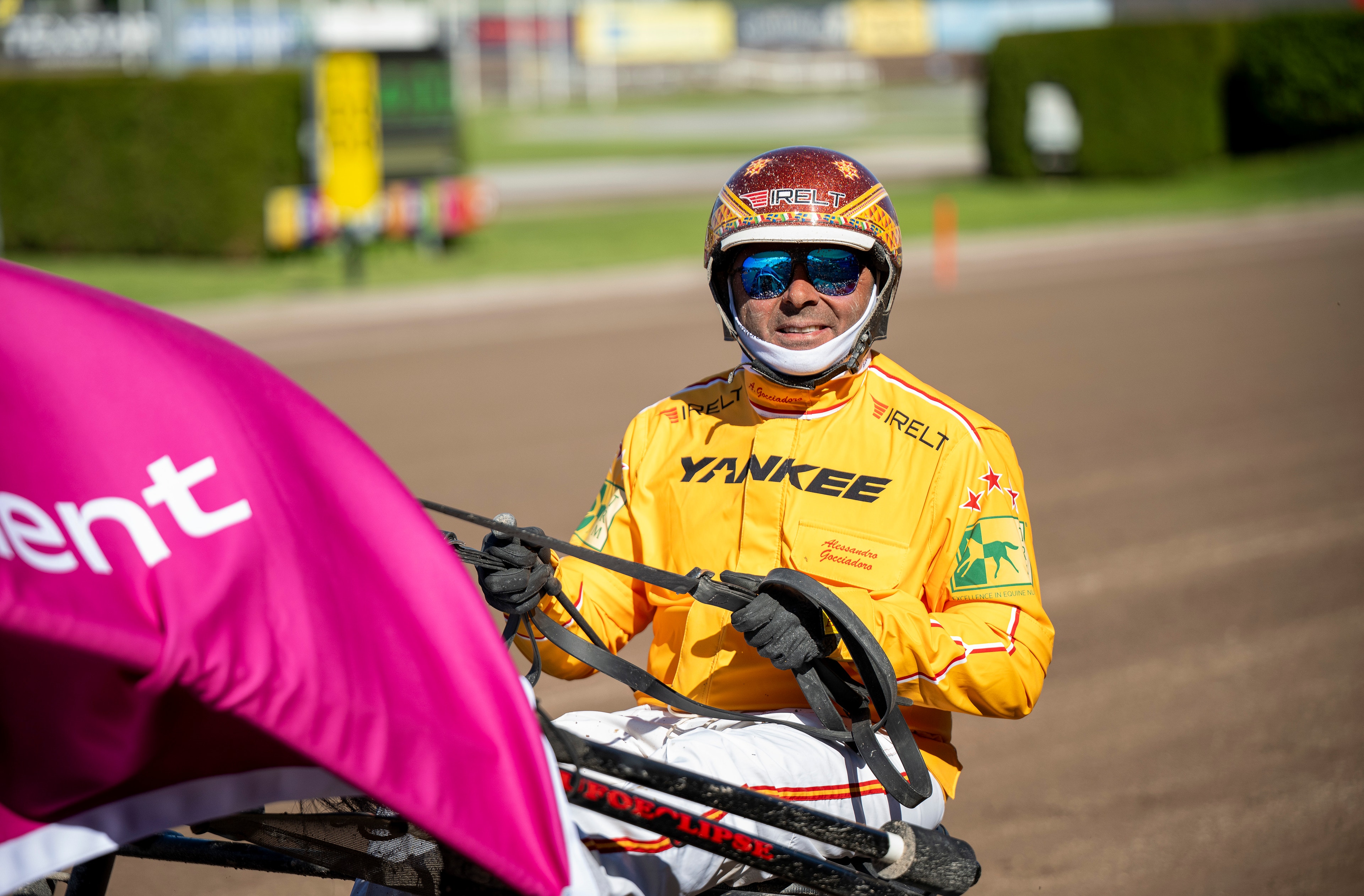 En kuskdragare i gul racingdräkt och hjälm sitter i en kuskdragarvagn på en racingbana.