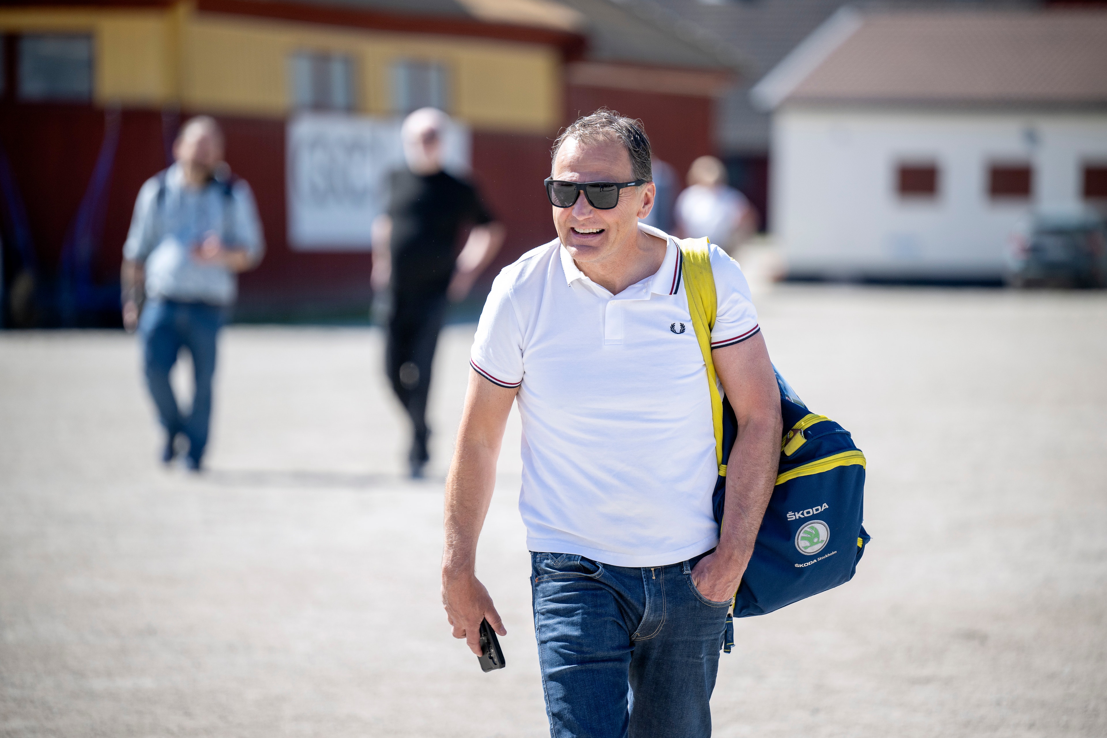 Man i vit t-shirt och jeans går på en gård med en blå väska över axeln. Andra personer och byggnader syns i bakgrunden.