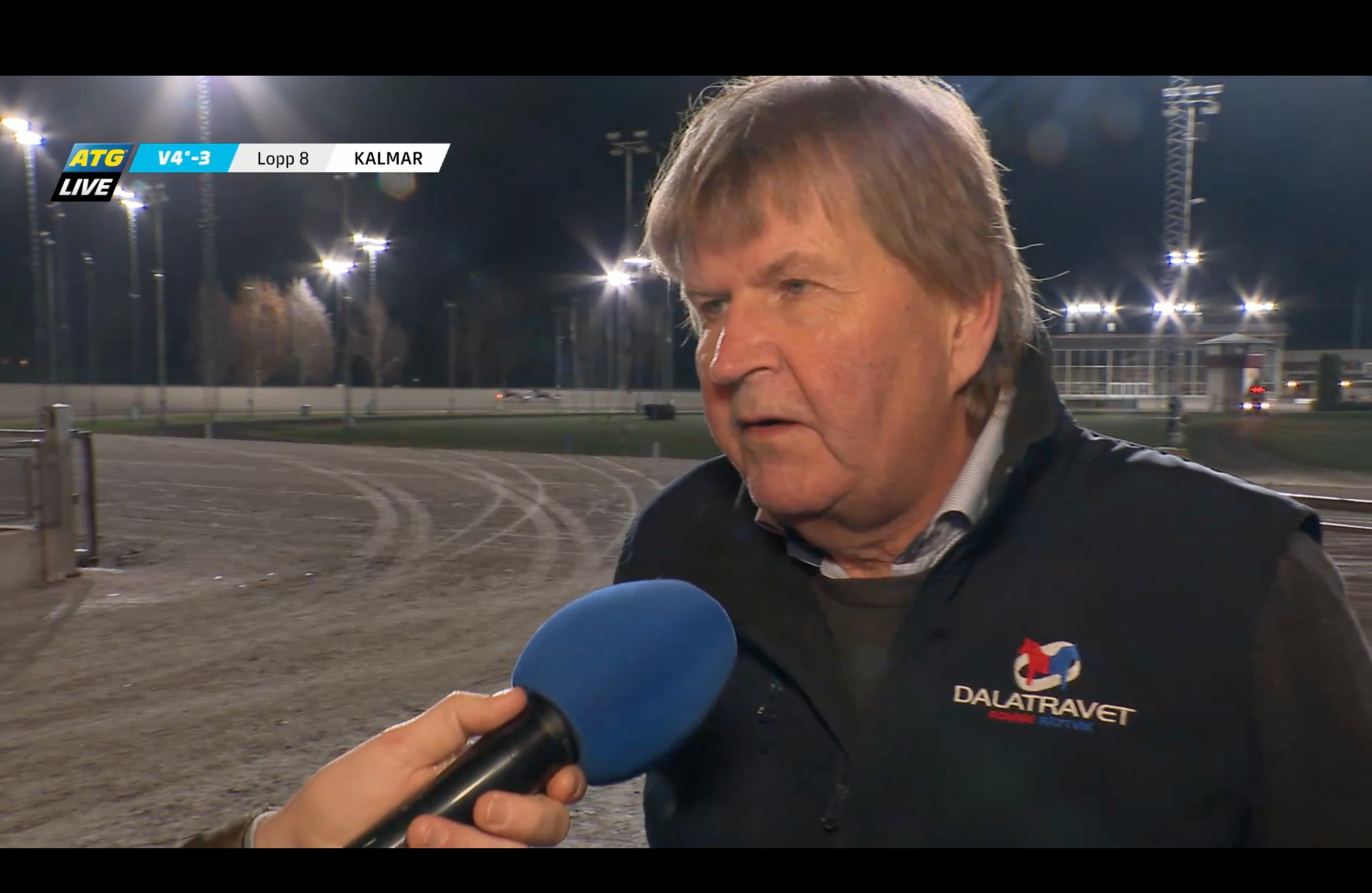 En äldre man med grått hår i svart väst intervjuas med en mikrofon framför en travbana i kvällsljus, i en tv-sändning från ATG Live.
