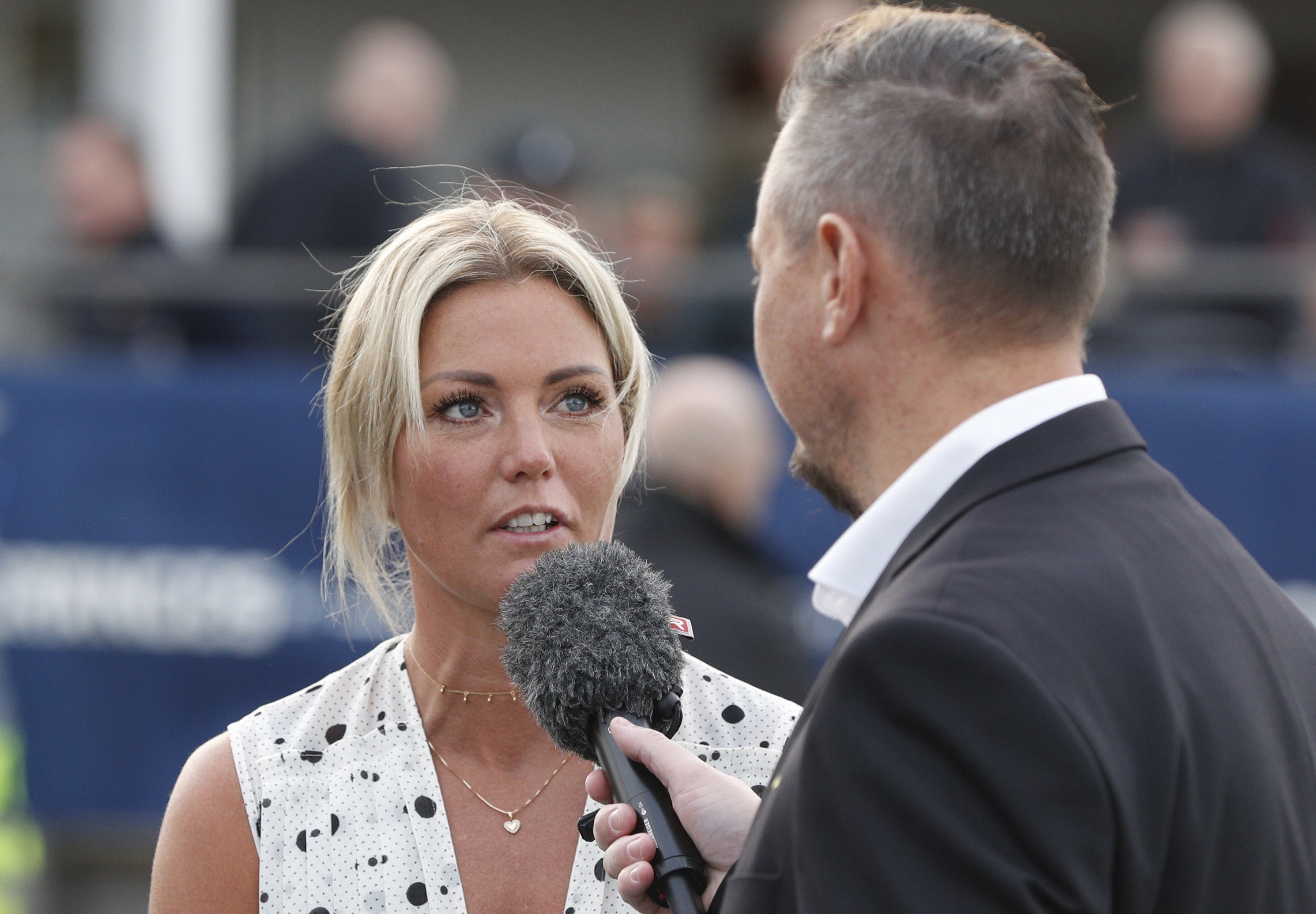 Två personer i en intervjusituation vid ett sportarrangemang. En kvinna med blont hår i en vit klänning med prickar intervjuas av en man i mörk kostym som håller en mikrofon.