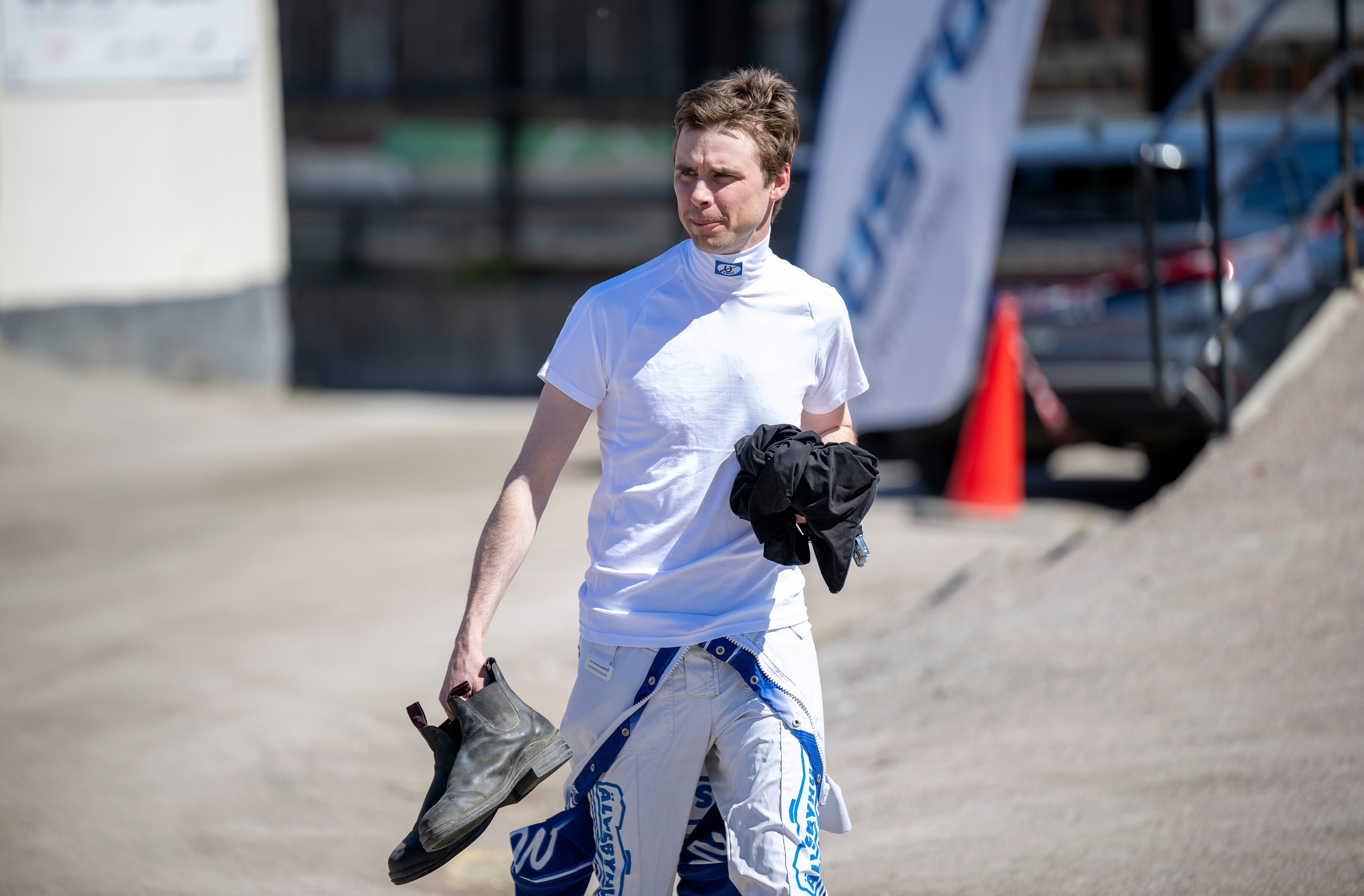 En person i vit t-shirt och blå motorsportbyxor står på en racerbana och håller motorsporthandskar i handen.