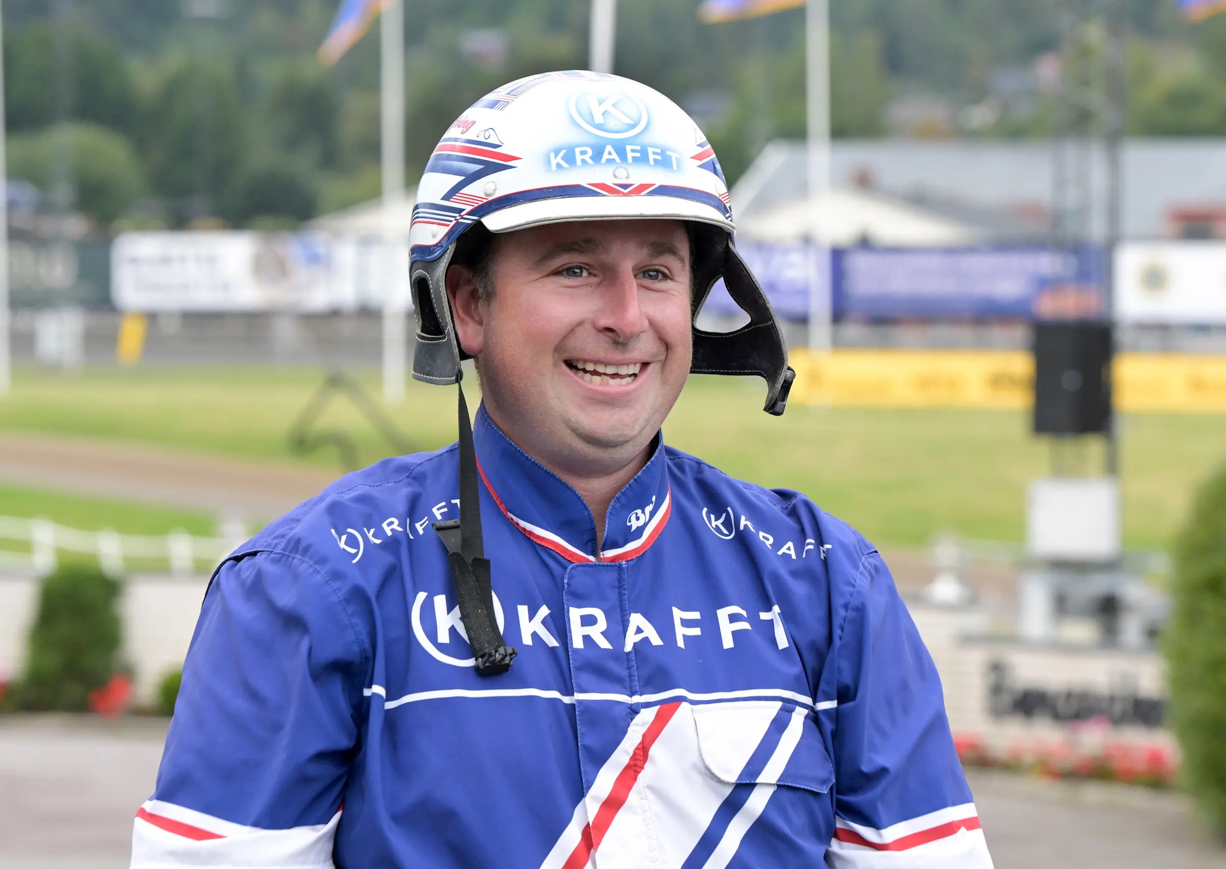Porträtt av en man i blå Krafftragg-dräkt och vit hjälm på en travbana.