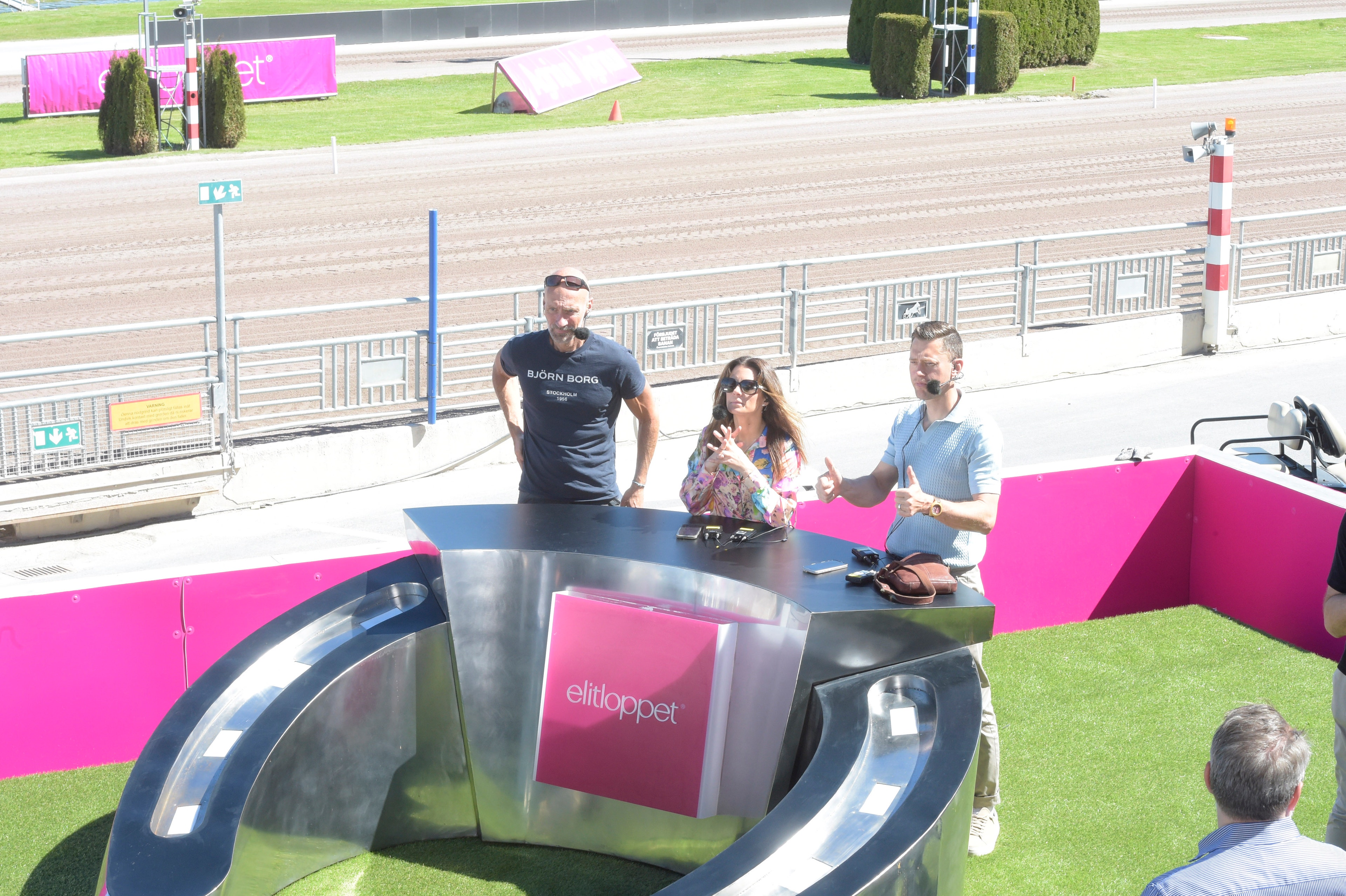 Utomhusevent vid en racerbana. Flera personer står vid en modern utställningsmonter med rosa och svart design. I bakgrunden syns racerbanan, gräsytor och racingutrustning.